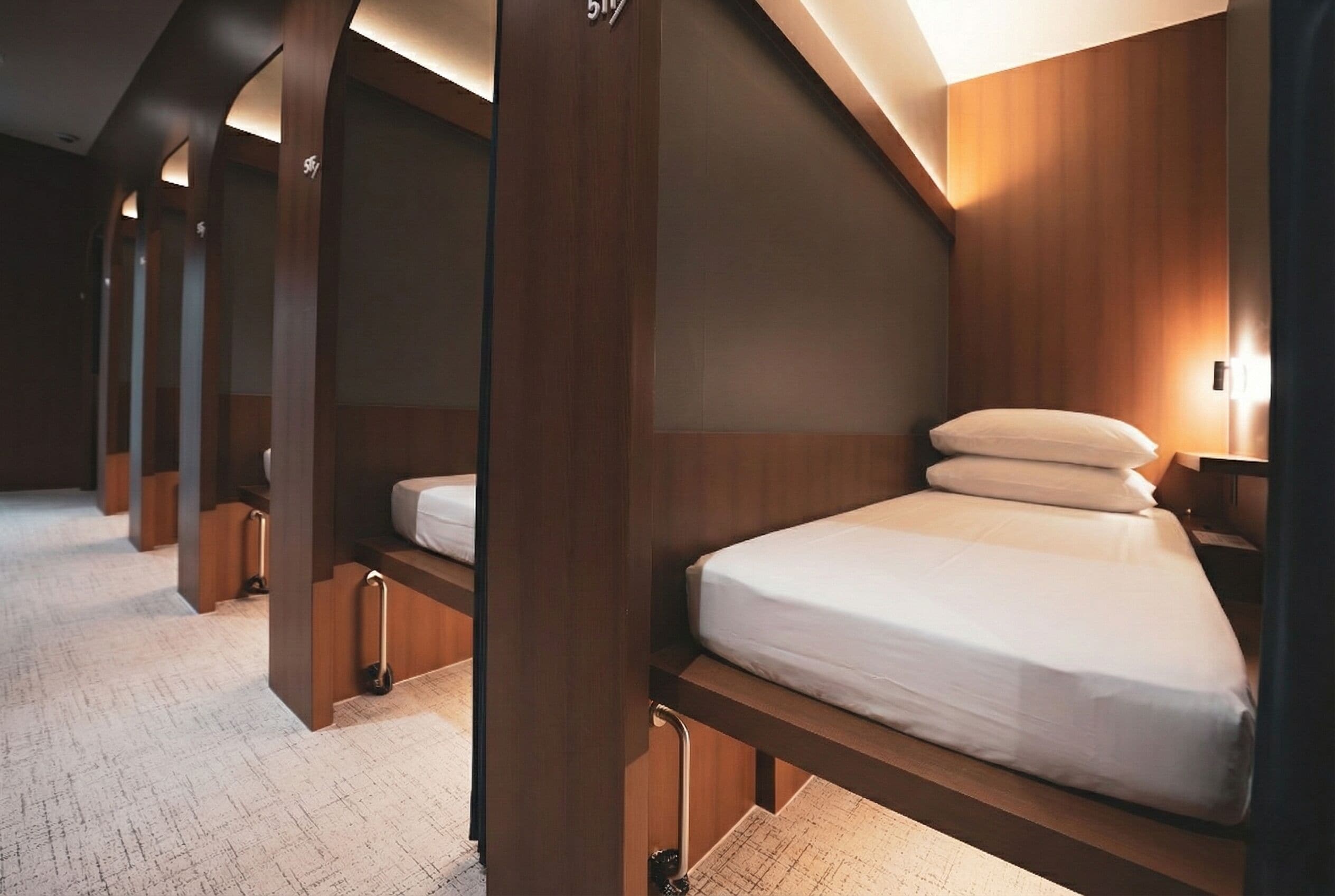 A row of wooden bunk beds with white bedding and headboards, each with a small bedside table and lamp.