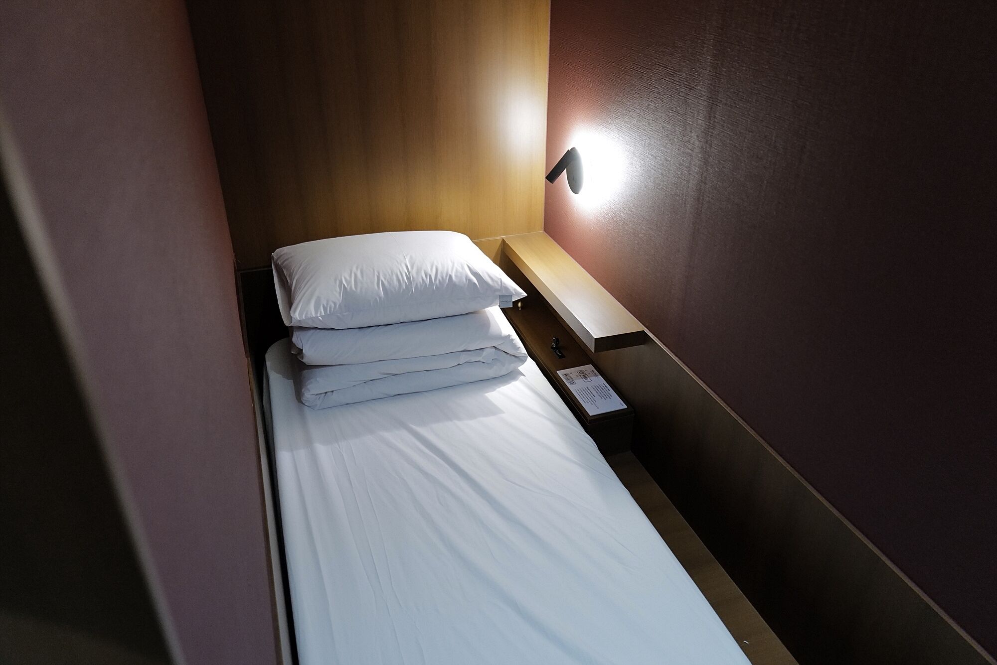 A narrow hotel room with a single bed, a nightstand, and a wall-mounted light.