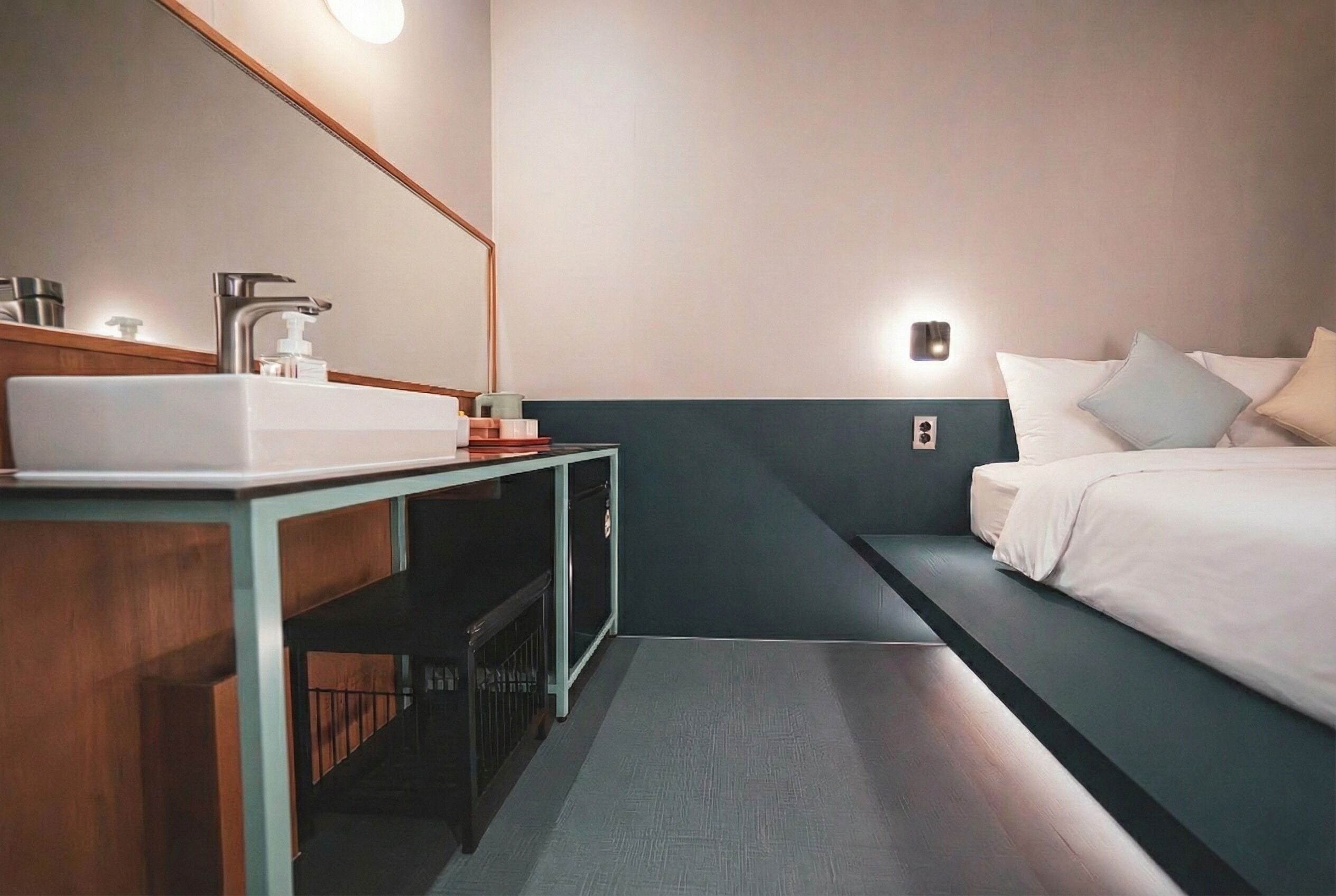 A modern hotel room with a wooden vanity, a large mirror, a white sink, a bedside table, a bed with white linens, and a small wall-mounted light.
