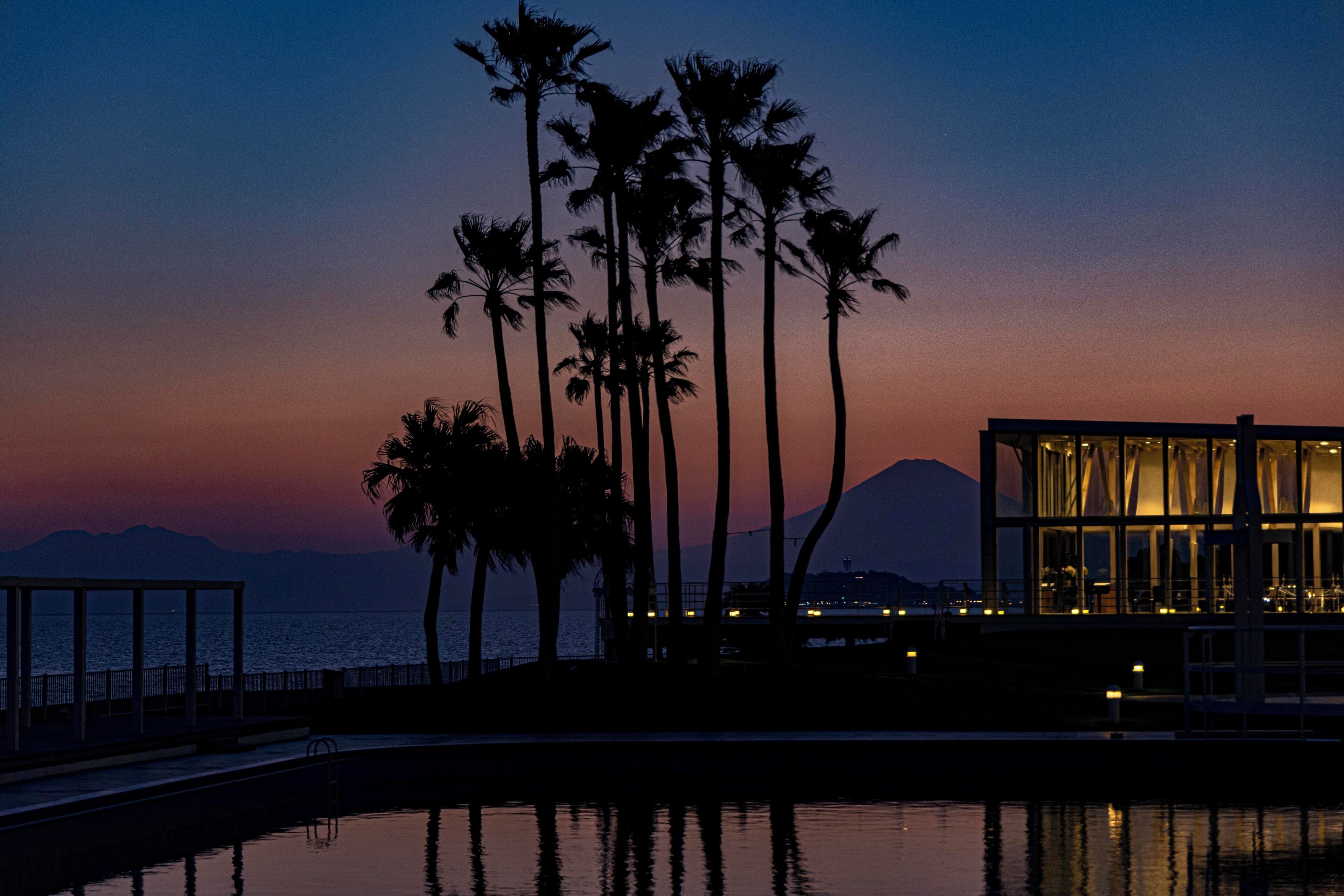 Un resort costero con piscina, palmeras y un edificio moderno con ventanas amplias.