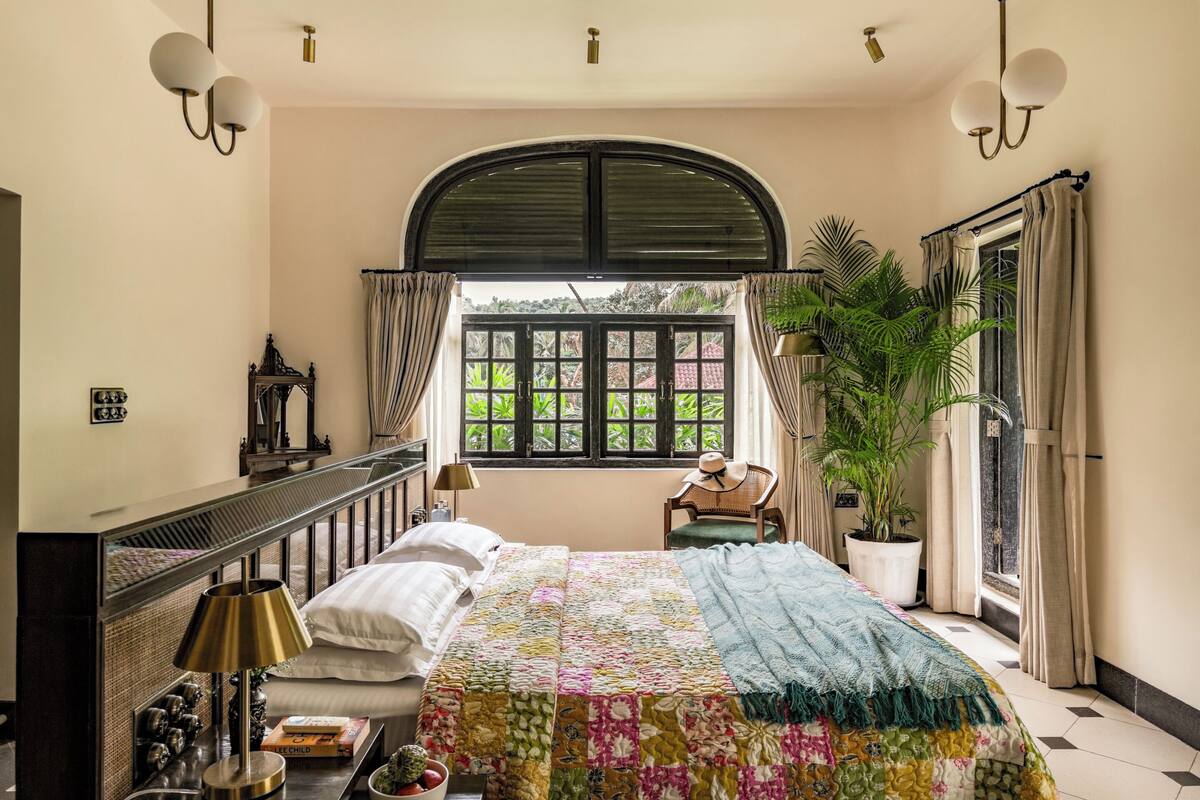 Ein Schlafzimmer mit einem großen Bett, einer Topfpflanze, einem Sessel und Blick durch ein Fenster.