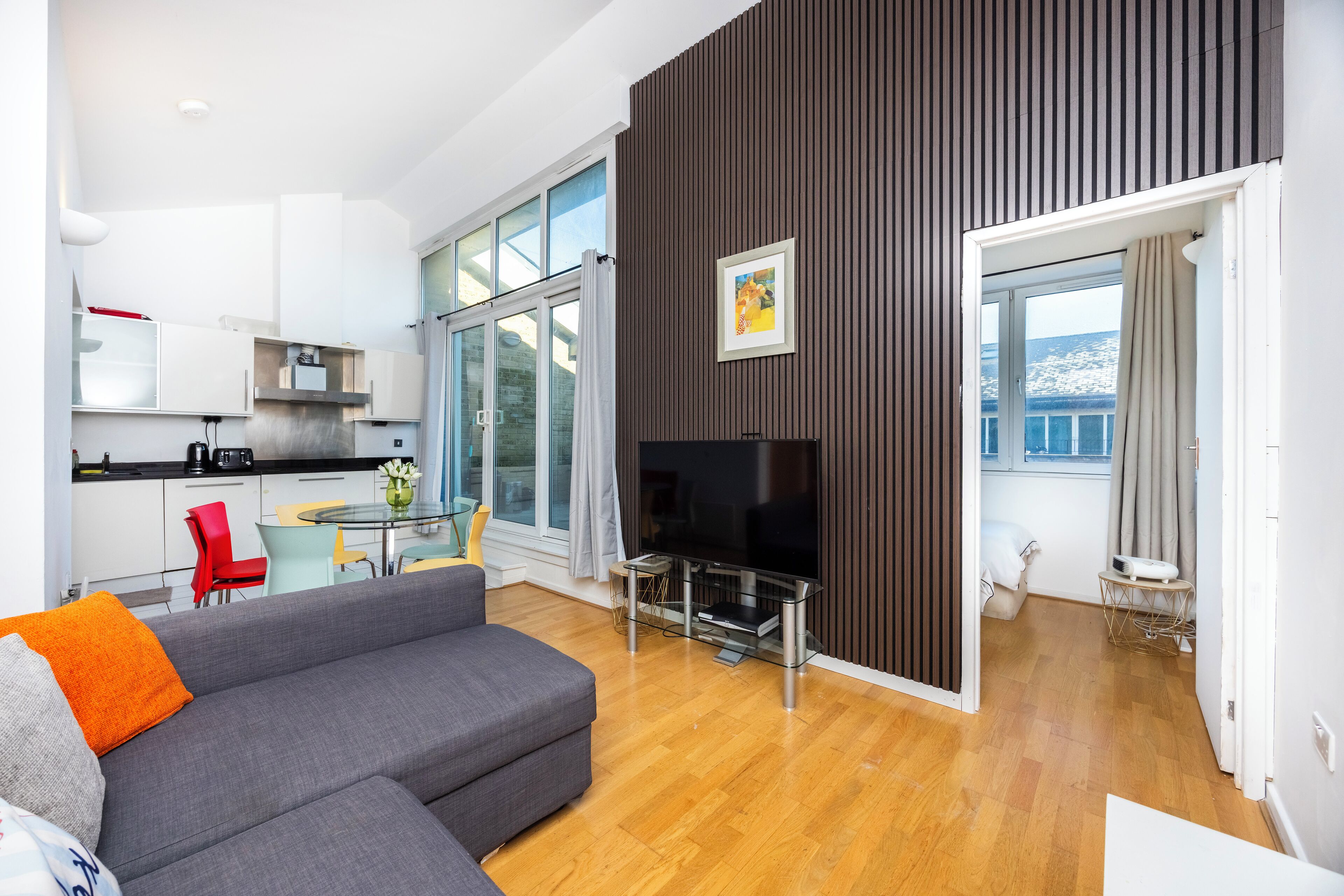 Een moderne woonkamer met een grijze bank, een flatscreen televisie en een eethoek met een glazen tafel en stoelen.