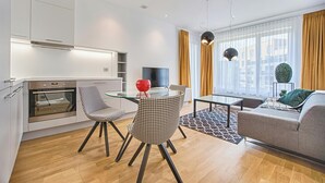 Een moderne keuken met oven, een eetgedeelte met een glazen tafel en stoelen, een bank en een groot raam met gordijnen.