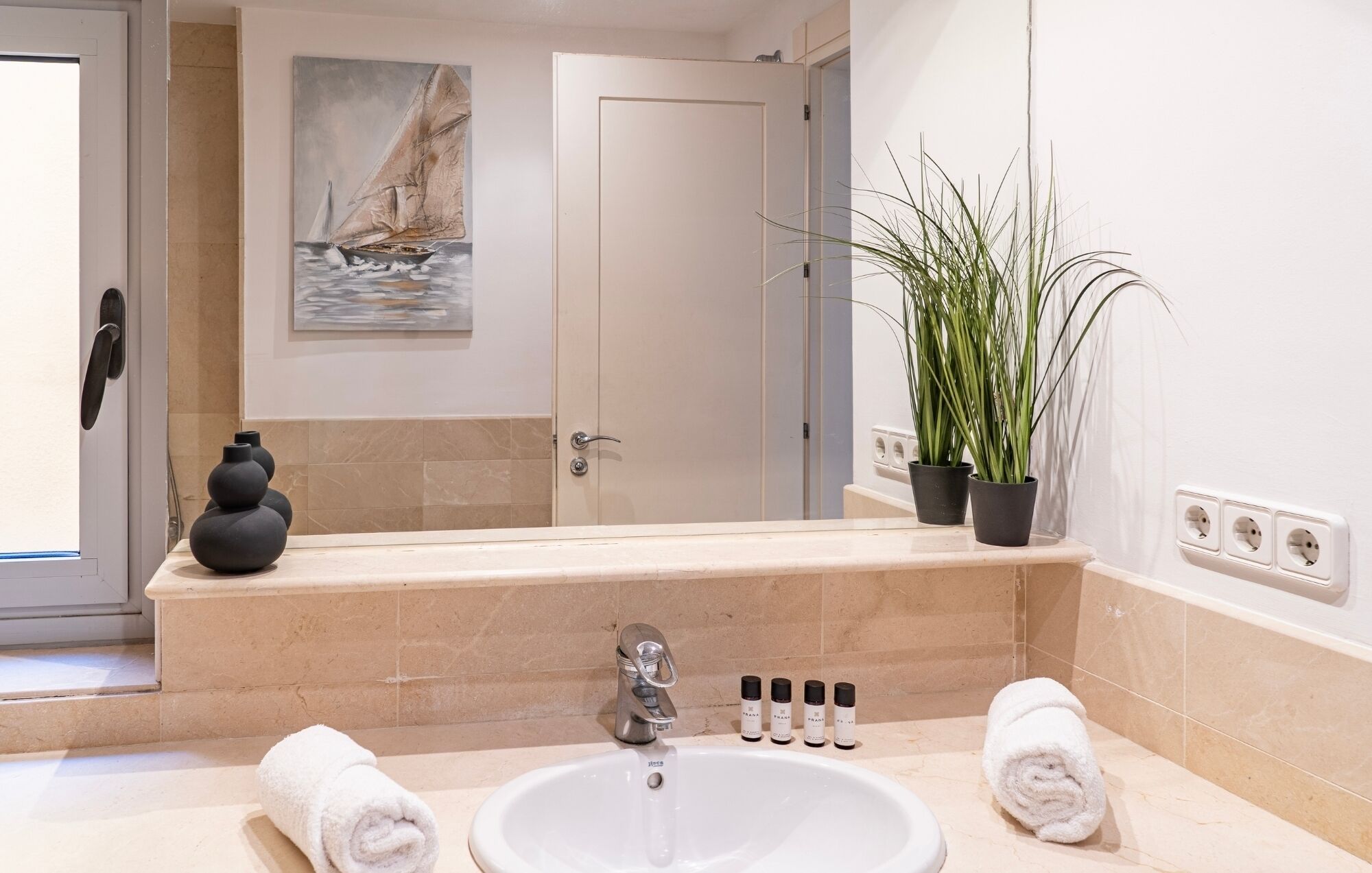 A bathroom with a large mirror, a sink, a towel, a potted plant, and a painting of a sailboat.