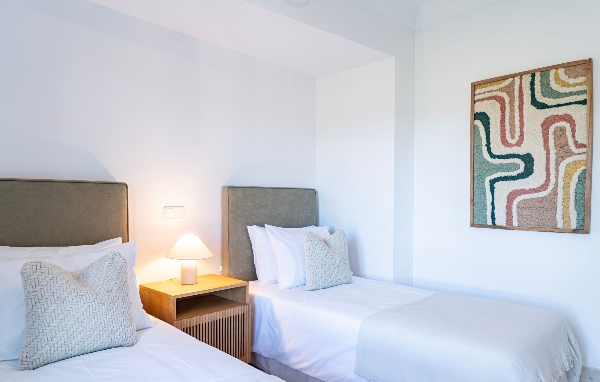 A hotel room with two beds, a nightstand with a lamp, and a colorful abstract painting on the wall.