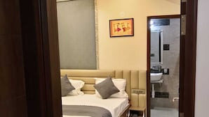 A modern bedroom with a bed, bedside tables, a nightstand, a mirror, and a bathroom visible through an open door. - Hotel Sky Inn (Sikar)