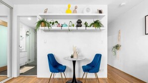 Een moderne kamer met een witte muur, twee blauwe stoelen, een klein rond tafeltje en planken met planten en decoratieve objecten.