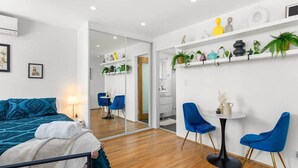 Een moderne hotelkamer met een bed, een bureau en stoelen. Er hangt een spiegel aan de muur, en er staan planten en decoratieve objecten op de planken.