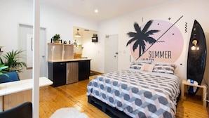 A compact bedroom with a bed, a small kitchenette, a desk, and a surfboard decoration. - Beachfront Bondi Studio with Aircon (North Bondi)