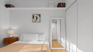 Een slaapkamer met een bed, een nachtkastje met een lamp, een lijst met kunst aan de muur en een boekenplank vol boeken.