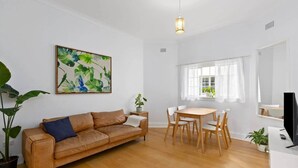 Een moderne woonkamer met een bruine leren bank, een eettafel met stoelen, een schilderij aan de muur en een televisie op een dressoir.