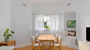 Een kamer met een houten eettafel, witte stoelen, een potplant, een raam met dunne gordijnen, een schilderij aan de muur en een televisie op een kast.