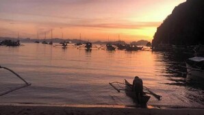 A coastal scene with boats docked along the shore, a sandy beach, and a rocky cliff. - Apoheo Beach Cabin (El Nido)