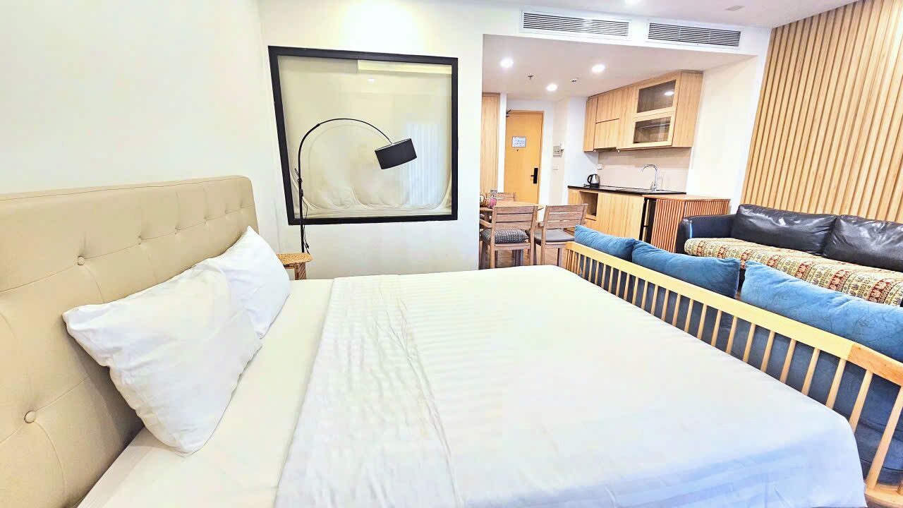 Une chambre d’hôtel avec un grand lit, un canapé, un téléviseur fixé au mur et, au fond, une petite cuisine.