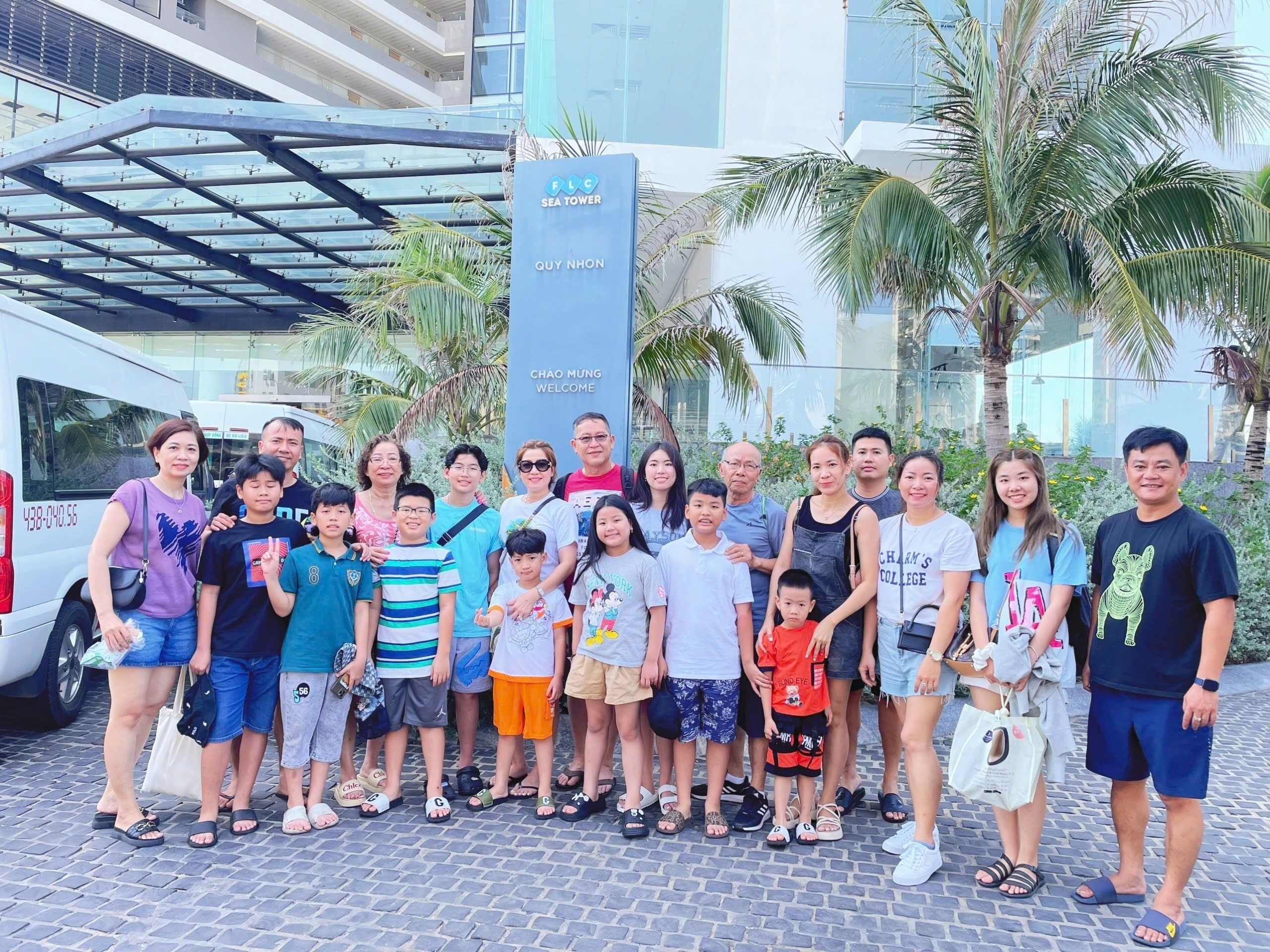 Un grupo de personas posando frente a un edificio moderno con un letrero que dice "FLC SEA TOWER QUY NHON CHAO MUNG WELCOME".