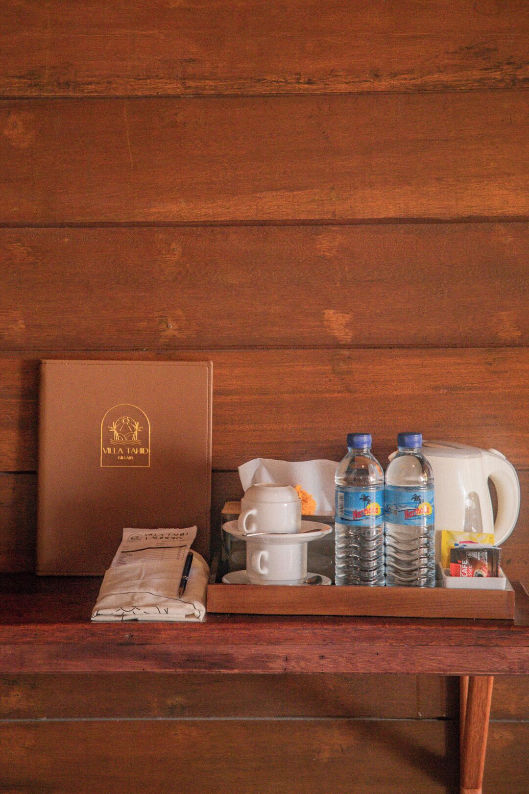 A wooden table with a brown leather-bound book, a white cup, two water bottles, a white electric kettle, a small packet, and a spoon.