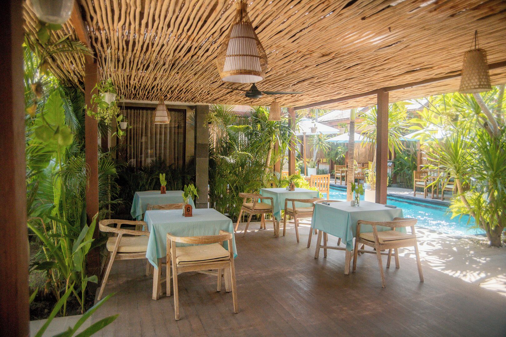 Un área de comedor al aire libre con mesas y sillas de madera, rodeada de exuberante vegetación y una piscina.