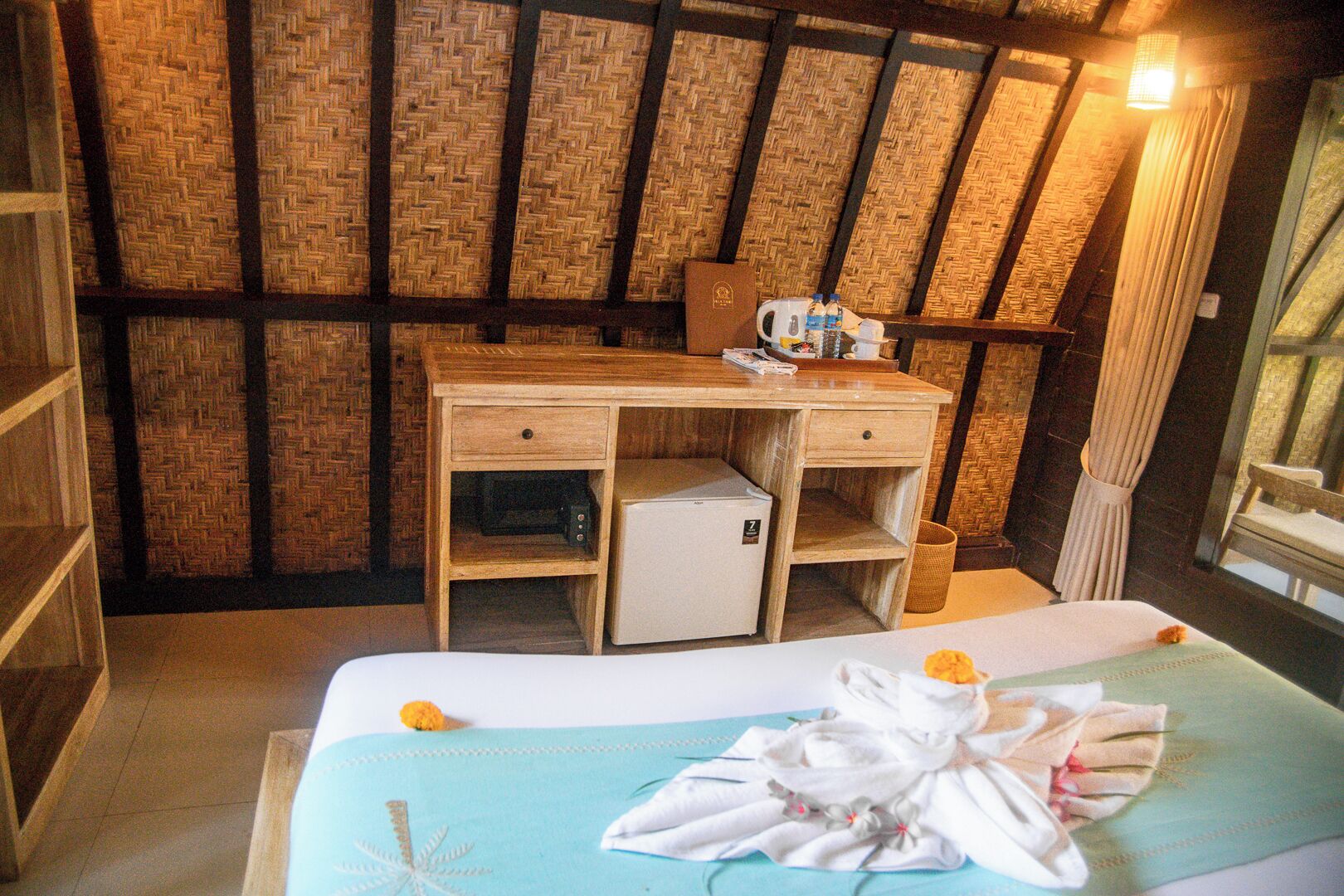 A room with a bed, a desk, a small refrigerator, and a woven wall.