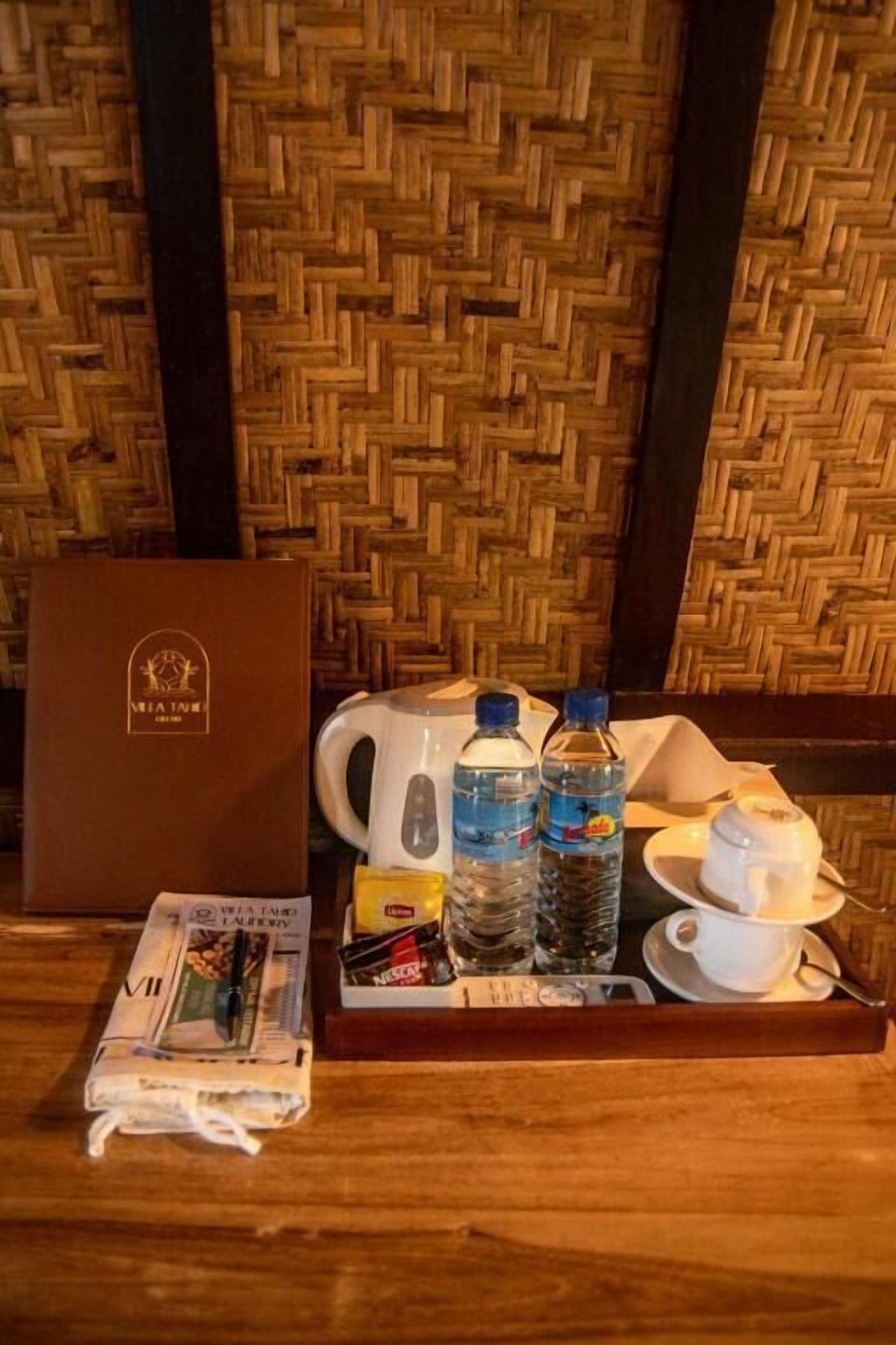 Ein Holztablett mit einer braunen Schachtel, einer Zeitung, einer Wasserflasche, einer Tasse und einer Kaffeetasse auf einem Tisch vor einem Hintergrund mit gewebter Wand.