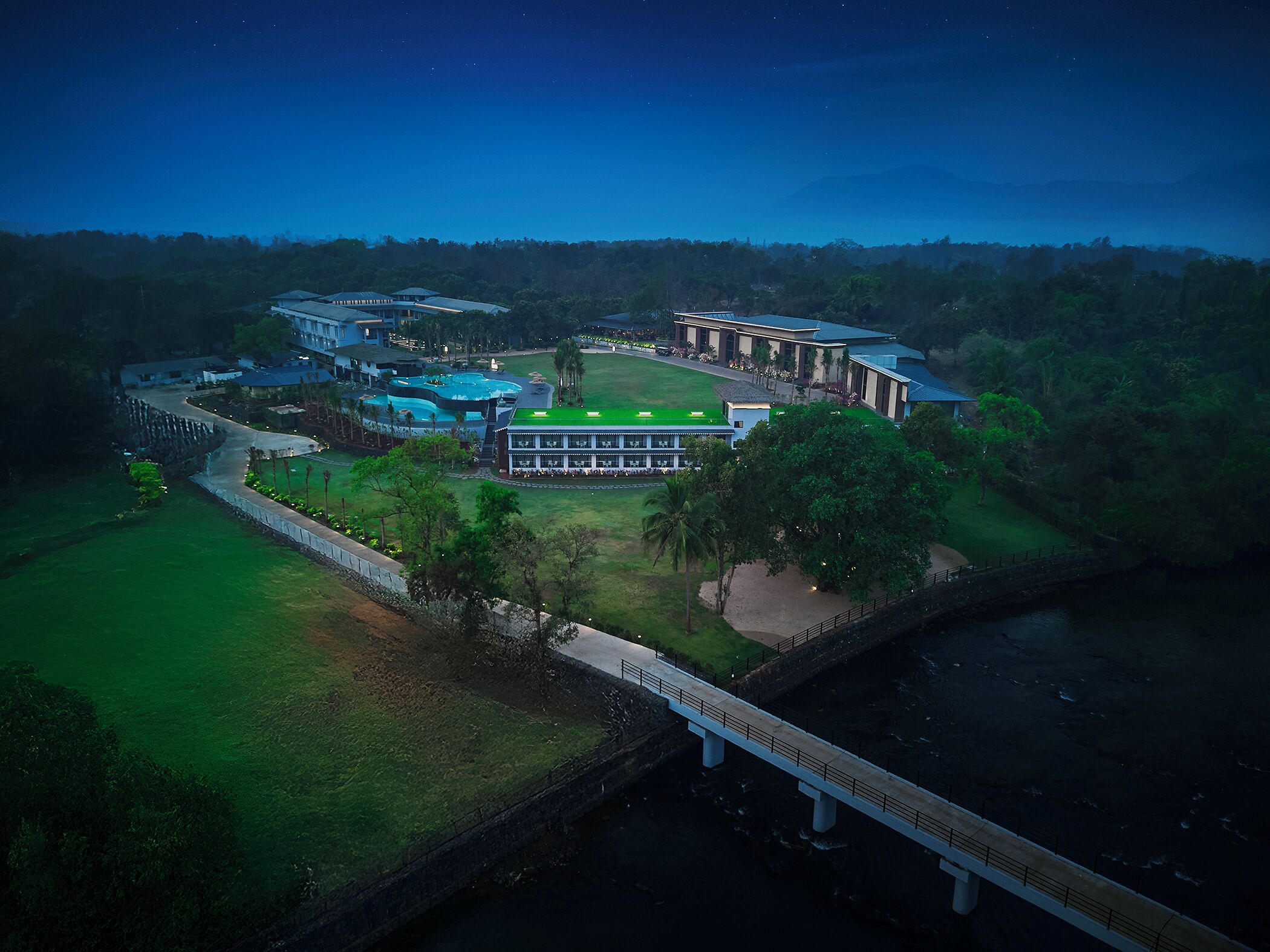 An aerial view of a resort with a swimming pool, a large building with a green roof, and a bridge over a body of water.