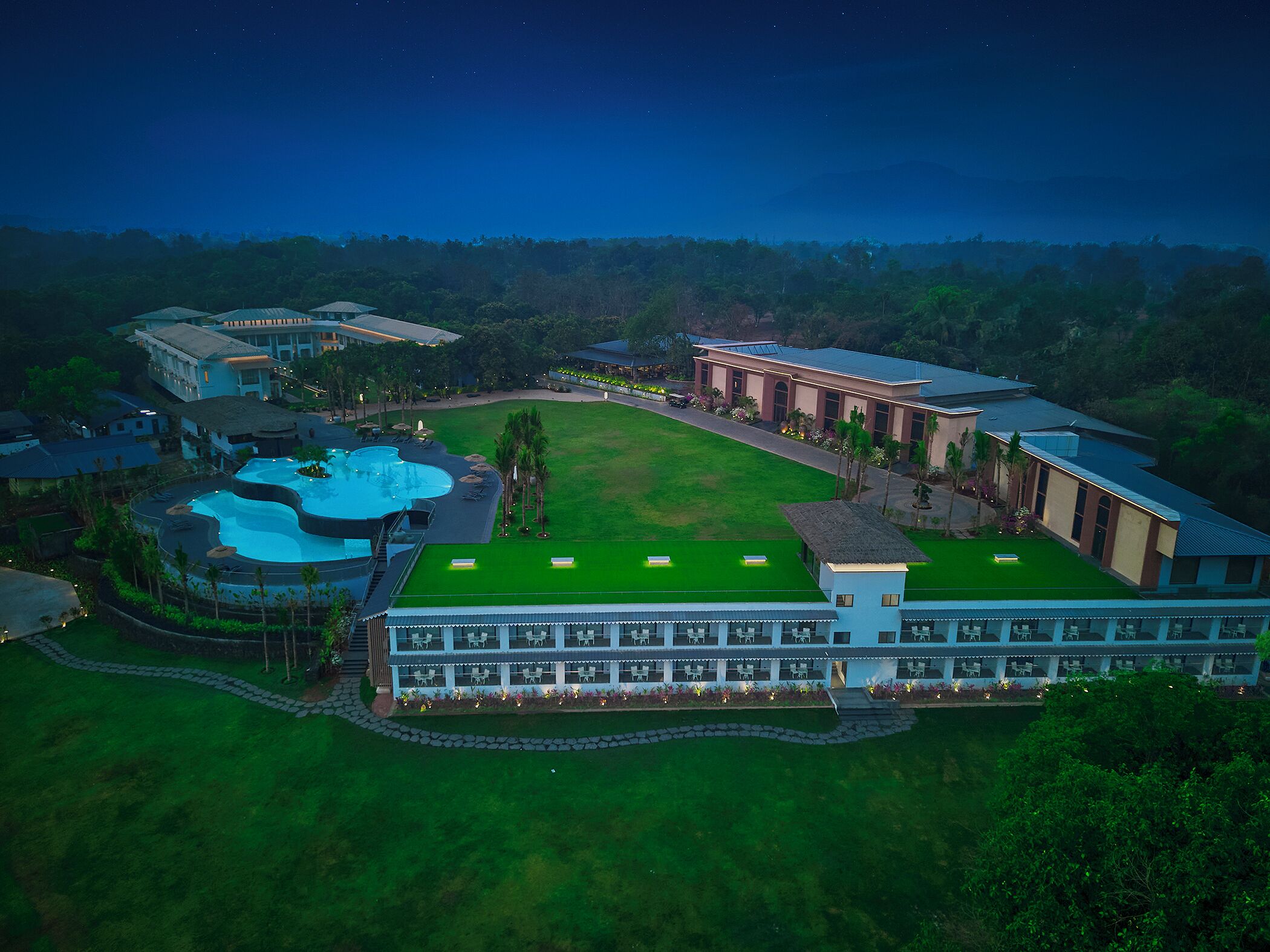 An aerial view of a resort with a large swimming pool, a tennis court, and multiple buildings with a green roof.