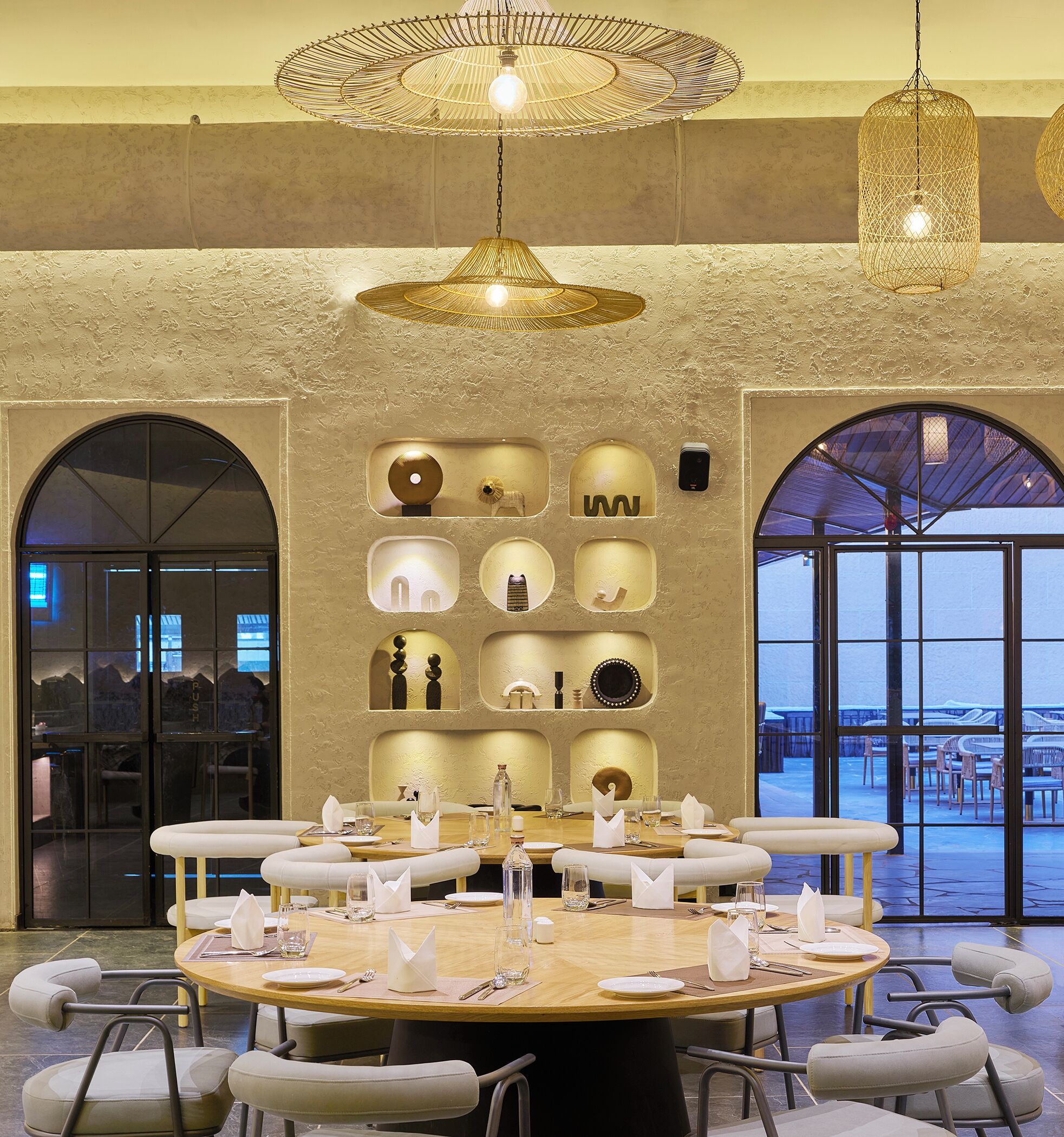 A dining area with a round table set for six, surrounded by cushioned chairs. There are decorative wall sconces and a large arched window with a view.