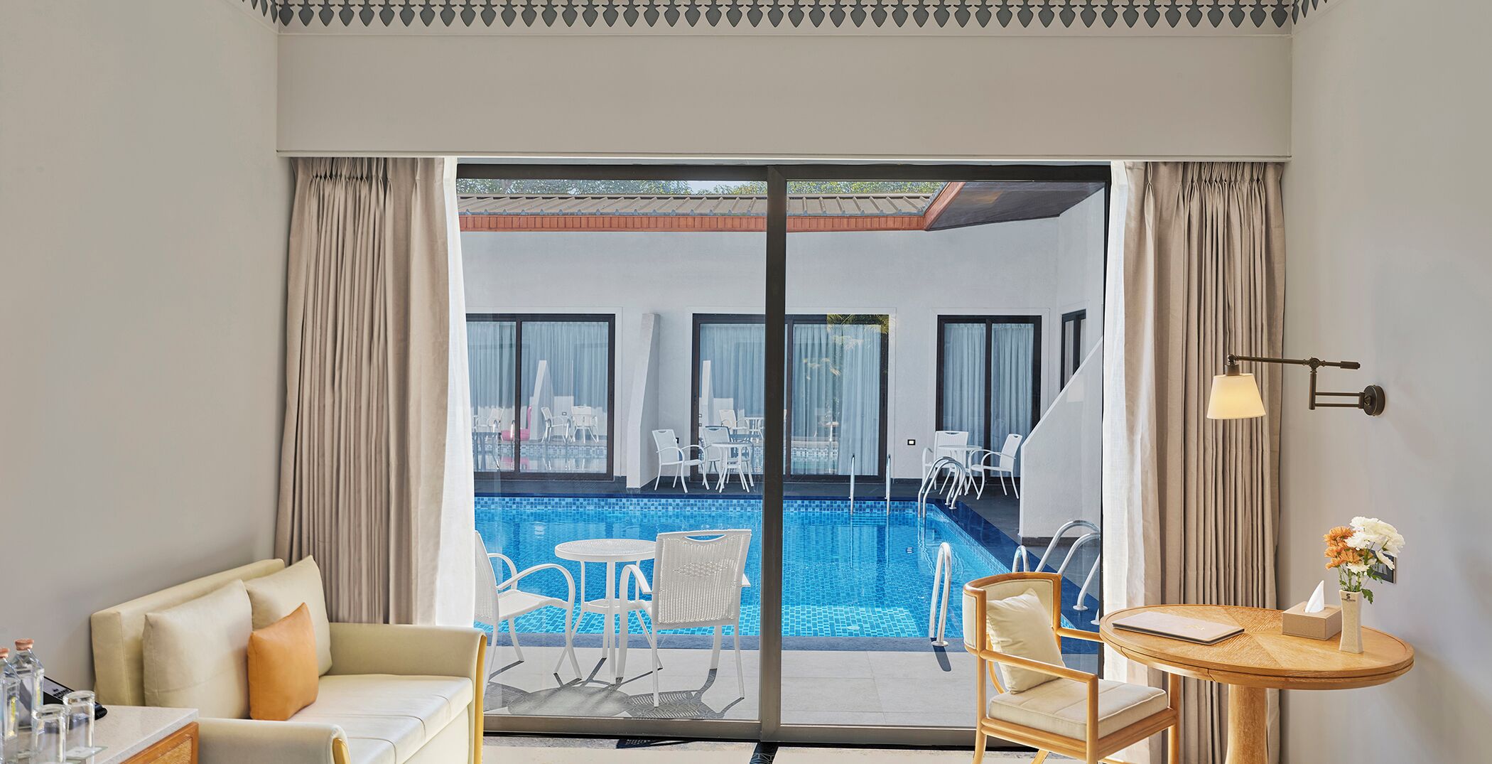 Ein Hotelzimmer mit einer großen Schiebeglasdoor, die zu einem Außenpoolbereich führt, mit weißen Stühlen und einem Tisch eingerichtet.
