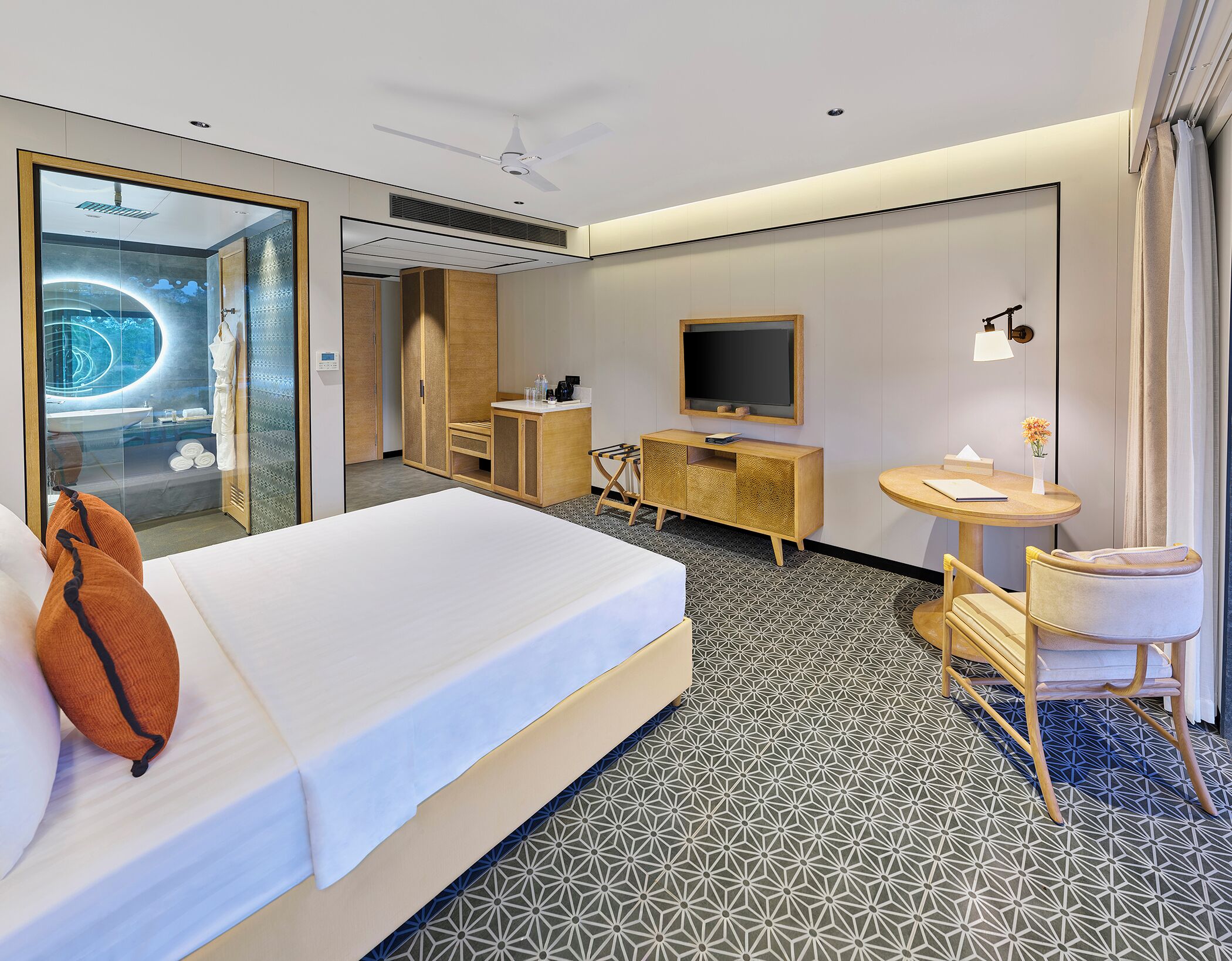 A hotel room with a bed, a desk, a TV, and a bathroom visible through a glass door.