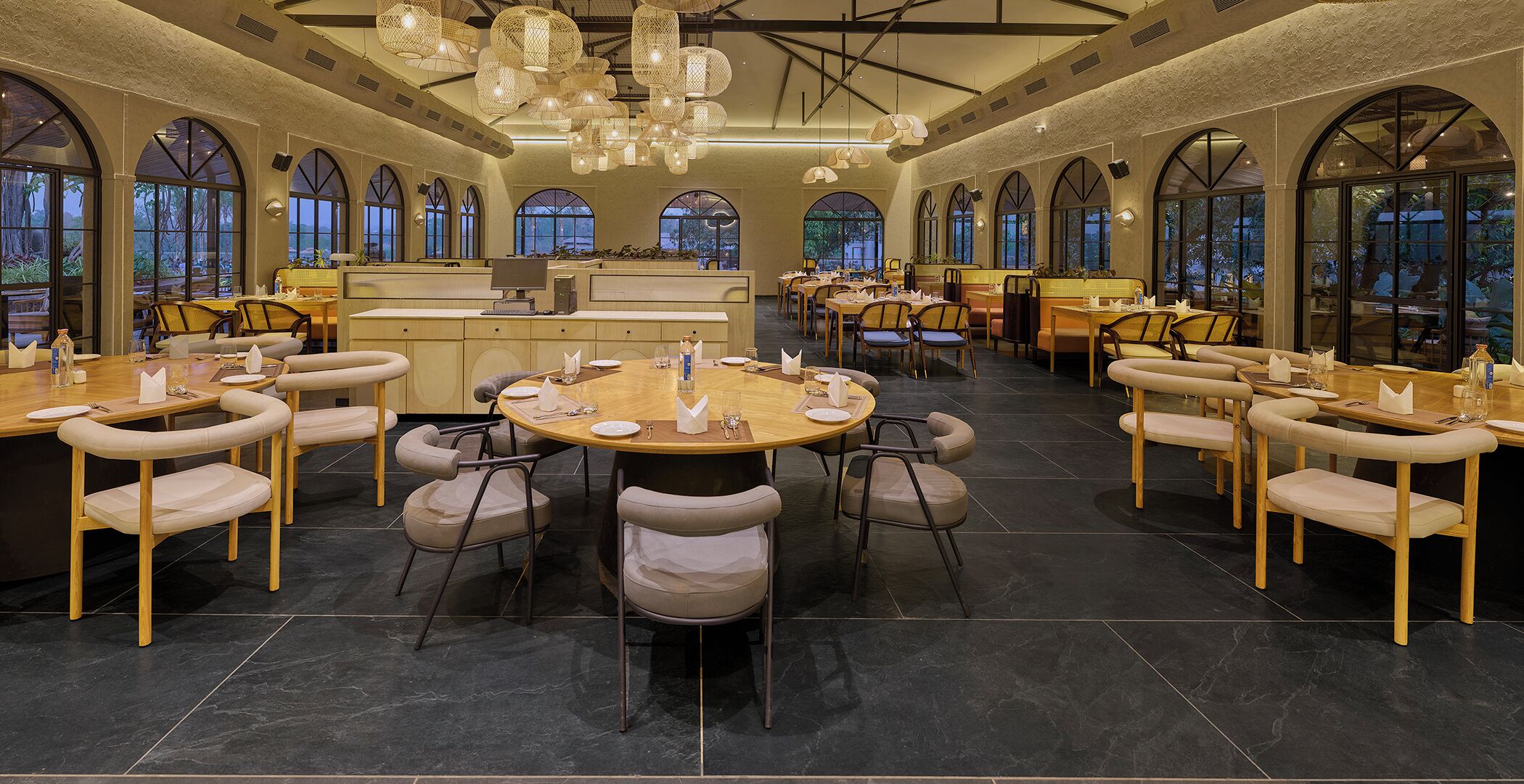 Een ruime eetruimte met ronde tafels en comfortabele stoelen, grote ramen en hangende lampen.