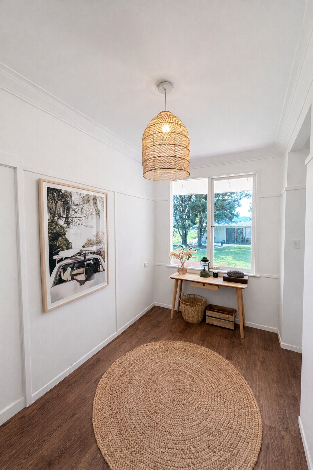 A room with a wooden floor, a round woven rug, a desk with a basket, a hanging light fixture, and a framed picture on the wall.