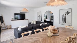 A modern living room with a large wooden dining table, black sofas, and a TV mounted on the wall.