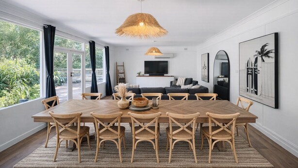A spacious dining area with a large wooden table and chairs, a woven pendant light, and a view of greenery through large windows.