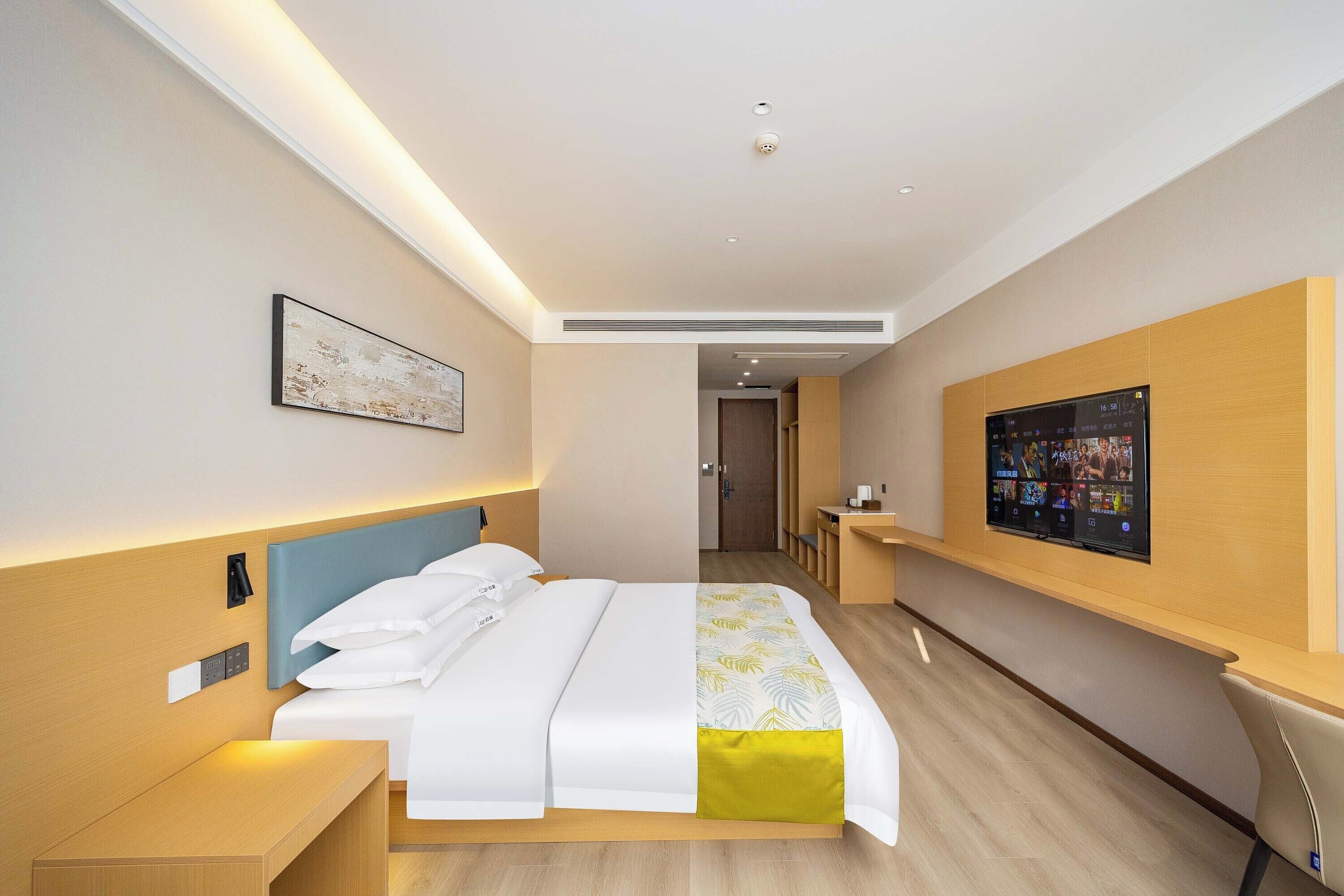 A modern hotel room with a neatly made bed, a bedside table, a wall-mounted TV, and a small seating area with a chair.