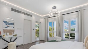 A modern bedroom with a large bed, a glass desk, a round mirror, and a view of the outdoors through large windows. - LUX The Serene Springs Villa 2 (Dubai)