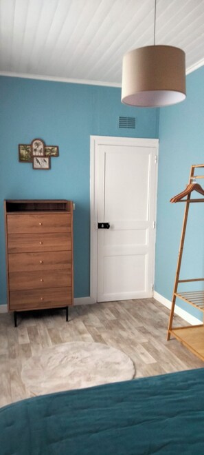 Quarto com cômoda de madeira, porta branca, luminária pendente e estante de madeira.