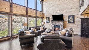 A spacious living room with a fireplace, large windows, and a flat-screen TV mounted above it. - Mountain View + Loft + Game Room + Hot Tub | Near Gatlinburg | Vixen Run Lodge (Gatlinburg)