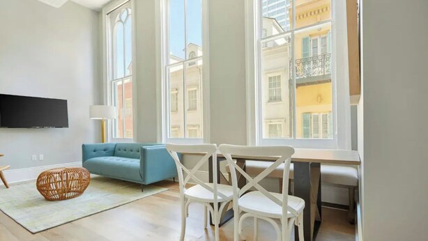 Ett modernt vardagsrum med en blå soffa, vita stolar, en platt-TV och stora fönster med utsikt över en stadsbild.