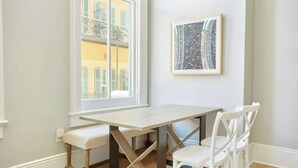 Ett rum med ett träbord och vita stolar, ett fönster med utsikt över byggnader och en inramad tavla på väggen.