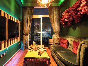 A modern living room with a checkered coffee table, a sofa with red cushions, a large flat-screen TV, and a view of a cityscape through a window. - Manzil - Stylish 1BR with Canal View (Dubai)