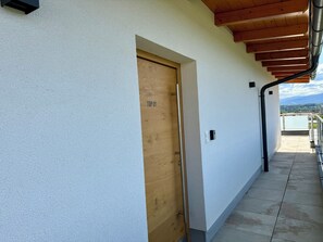 Ein Korridor mit einer Holztür mit der Aufschrift 'TOP 01', einem Metallgeländer und Blick auf einen Balkon mit blauem Himmel.