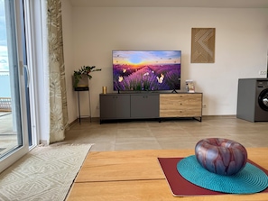 Ein Wohnzimmer mit Flachbildfernseher, Schrank, Waschmaschine, Wanddeko und einem Fenster mit Vorhängen.