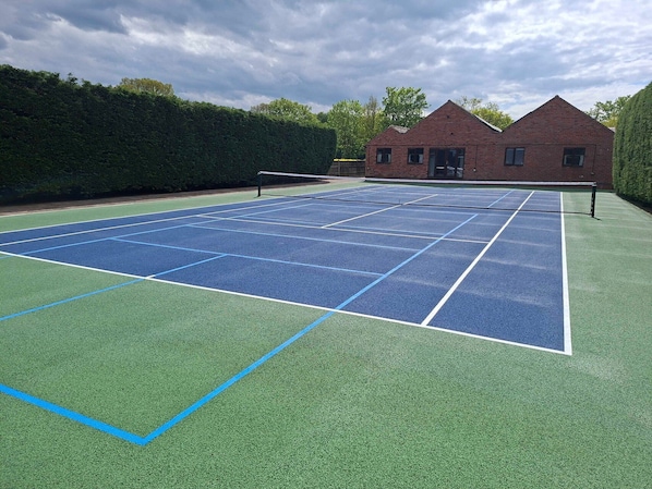 A tennis court with a net, surrounded by a hedge and a brick building.