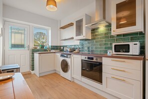 A compact kitchen with a washing machine, oven, microwave, and a window with a view of trees. - Pools Cottage 3 Billards Room-Games & Sports Court (Warwickshire)