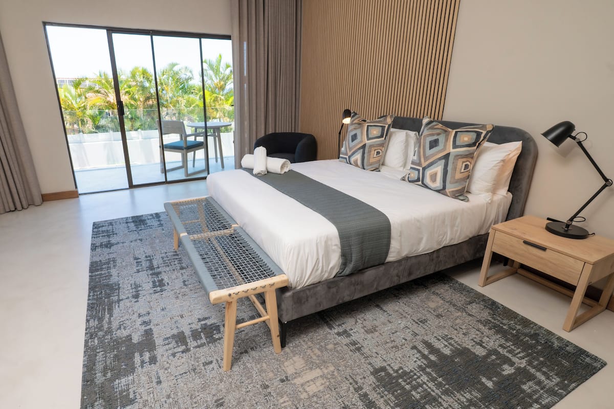 A modern bedroom with a large bed, a small desk, and a view of palm trees through the balcony doors.