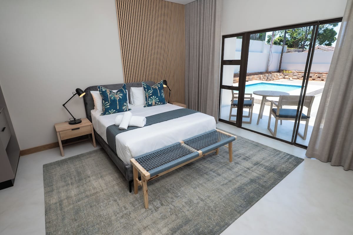 A modern bedroom with a large bed, a small bedside table, a bench, and a view of a swimming pool through a sliding glass door.