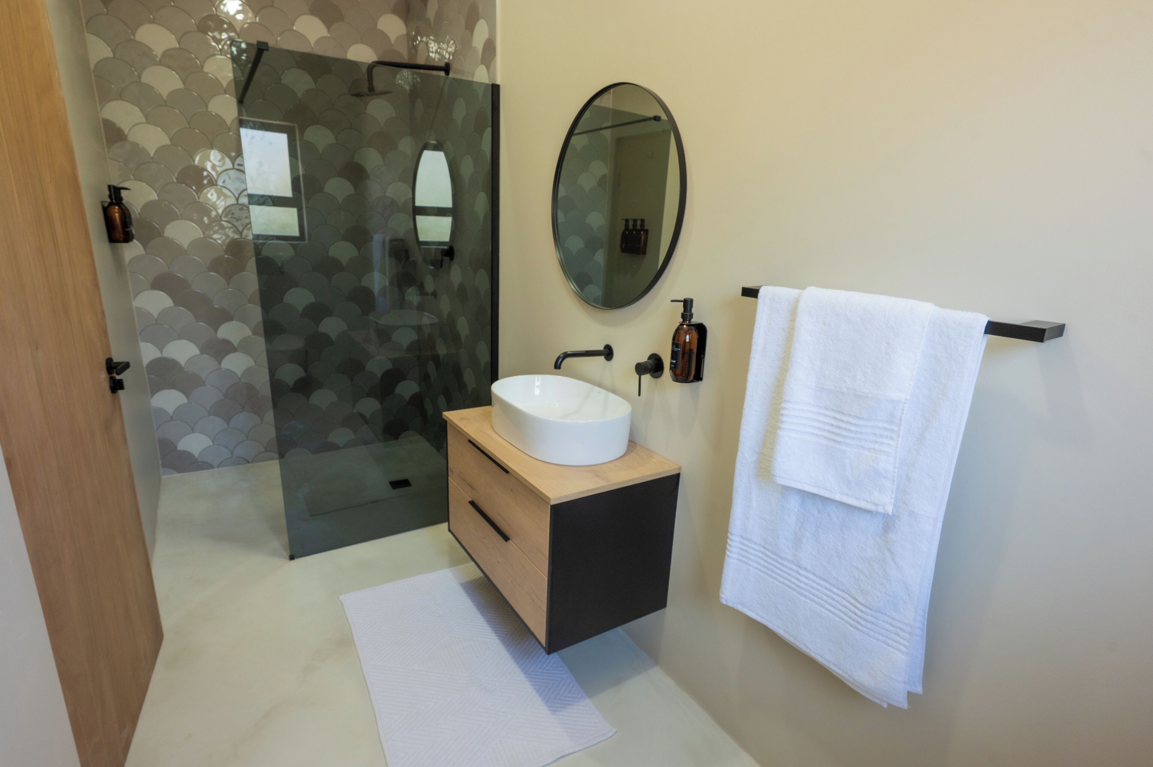 Un baño moderno con cabina de ducha de vidrio, tocador de madera con lavabo blanco, espejo redondo y perchero.