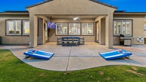 A covered patio area with two lounge chairs, a bench, and a television screen. - Terra Sol: Pool, Spa & Lakefront Serenity (Indio)