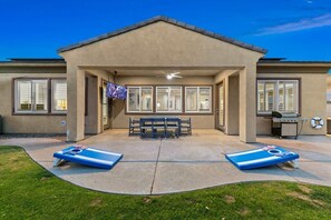 A covered patio area with two lounge chairs, a bench, and a television screen. - Terra Sol: Pool, Spa & Lakefront Serenity (Indio)