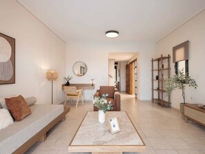 Ein modernes Wohnzimmer mit einer Couch, einem Couchtisch und einem Schreibtisch mit Stuhl. Ein Bücherregal und ein Blumentopf sind ebenfalls vorhanden.