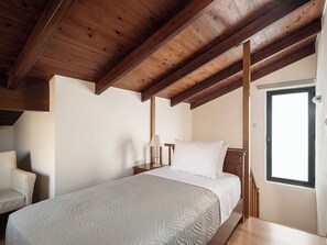 Ein Schlafzimmer mit Holzbalkendecke, einem Bett mit weißer Bettdecke, einem Nachttisch mit Lampe und einem Fenster.